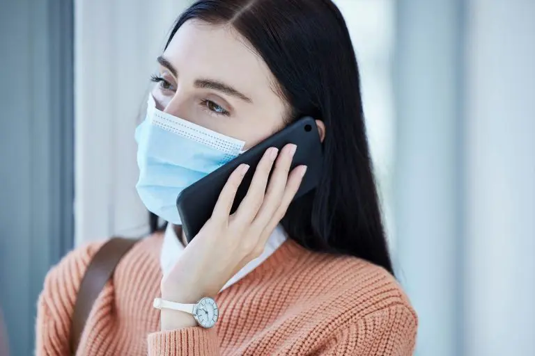 Woman, covid and mask on a phone call by window looking out at home for safety and quarantine indoo