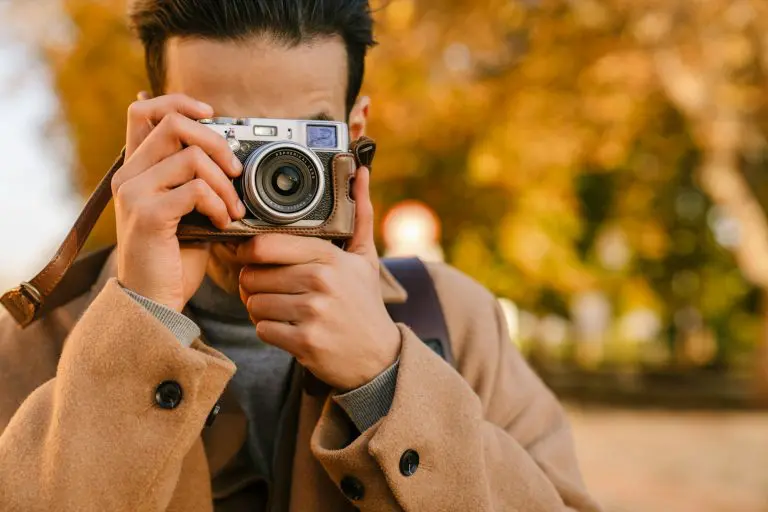 Young man wearing coat taking photo on retro camera in autumn park