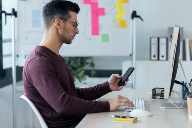 Handsome entrepreneur sending messages with smart phone while working with computer